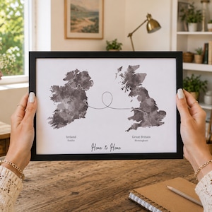 Op de afbeelding: Een ingelijste print met een grijze aquarelkaart van Ierland en Groot-Brittannië op een witte achtergrond. Een dunne zwarte lijn verbindt de twee landmassa's. De tekst onderaan luidt Ireland Dublin, Great Britain Birmingham en Home to Home.