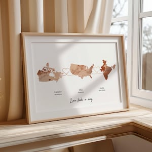 May include: Framed print featuring watercolor maps of Canada, the USA, and India, connected by heart-shaped lines. The print includes the text "Love finds a way" and city names. The frame is light-colored wood.