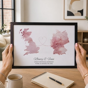 Peut inclure: Une impression encadrée présentant des cartes aquarelles de la Grande-Bretagne et de l'Allemagne en vieux rose, reliées par une ligne fine formant un cœur. Le texte indique : Nancy & Finn, GREAT BRITAIN & GERMANY, HOME IS WHERE WE ARE.