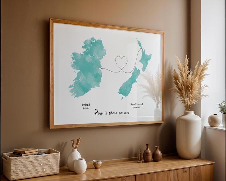 May include: A framed print featuring watercolor maps of Ireland and New Zealand, connected by a heart-shaped line. The text "Home is where we are" is written below the maps. The frame is made of light-colored wood.