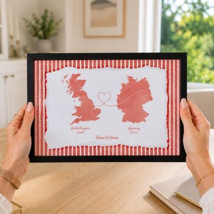 Könnte beinhalten: Ein gerahmter Druck mit einer roten Aquarellkarte von Großbritannien und Deutschland auf weißem Hintergrund mit abgerissenen Kanten. Eine dünne rote Linie verbindet beide Länder mit einem Herz in der Mitte. Der Text lautet United Kingdom, London, Germany, Berlin und Home to Home.