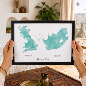 May include: A framed print featuring a teal watercolor map of Great Britain and South Africa. A dotted line connects the two regions with a heart shape in the center. The text reads Great Britain, London, South Africa, Johannesburg, and Home to Home.