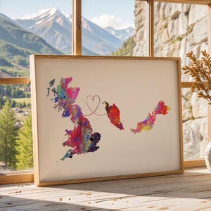 May include: A framed watercolour print showing the map outlines of the UK and Malaysia, linked by a heart-shaped line. The maps are filled with vibrant colours including pink, blue, yellow, and red, set against a cream background. The frame is made of wood.