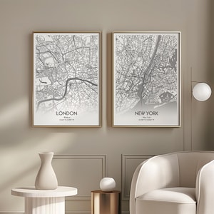 May include: Two framed city map prints. One shows London, the other New York, both in grayscale. The maps are detailed with streets and landmarks, set against a white background. The frames are gold-colored, and the prints are displayed on a neutral-toned wall.