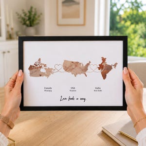 Può includere: Una stampa incorniciata su sfondo bianco con mappe in stile acquerello di Canada, USA e India in tonalità marroni. Linee sottili collegano i paesi con forme a cuore. Il testo recita: Canada Winnipeg, USA Houston, India New Delhi, Love finds a way.