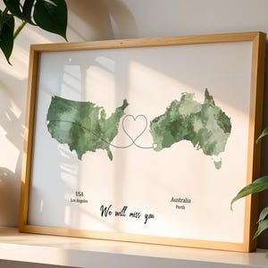 May include: A framed print featuring watercolor maps of the USA and Australia, connected by a heart-shaped design. The text "We will miss you" is below the maps, with city names. The frame is made of light-colored wood.