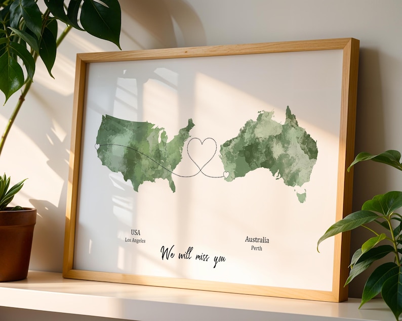 May include: A framed print featuring watercolor maps of the USA and Australia, connected by a heart-shaped design. The text "We will miss you" is below the maps, with city names. The frame is made of light-colored wood.