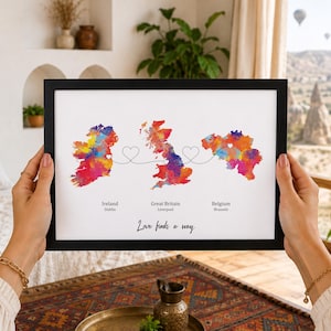 Può includere: Una stampa incorniciata con mappe in stile acquerello di Irlanda, Gran Bretagna e Belgio. Linee tratteggiate con cuori collegano i paesi. Il testo sottostante recita: Ireland Dublin, Great Britain Liverpool, Belgium Brussels e Love finds a way.