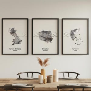 May include: Three framed prints with black frames, each showing a watercolour map of Great Britain, Poland, and Greece. The prints include the country name and capital city. A heart design connects the maps. The prints are displayed above a wooden table.