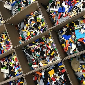 May include: Multiple cardboard boxes filled with colorful plastic building blocks. The boxes are arranged in a grid pattern, with some boxes partially open to reveal the contents.