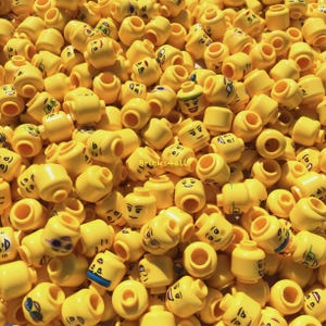 Könnte beinhalten: Eine große Ansammlung gelber Kunststoff-Spielzeugköpfe für Minifiguren, die jeweils einen einzigartigen aufgedruckten Gesichtsausdruck wie Lächeln, Zwinkern oder Brillen zeigen, mit dem Text Bricks4all in der Mitte des Haufens.
