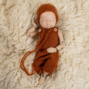 May include: A newborn photography prop featuring a doll wearing a rust-colored knitted romper and bonnet. The doll is lying on a cream-colored, fluffy rug. The romper has wooden button closures and a long, dangling tie.