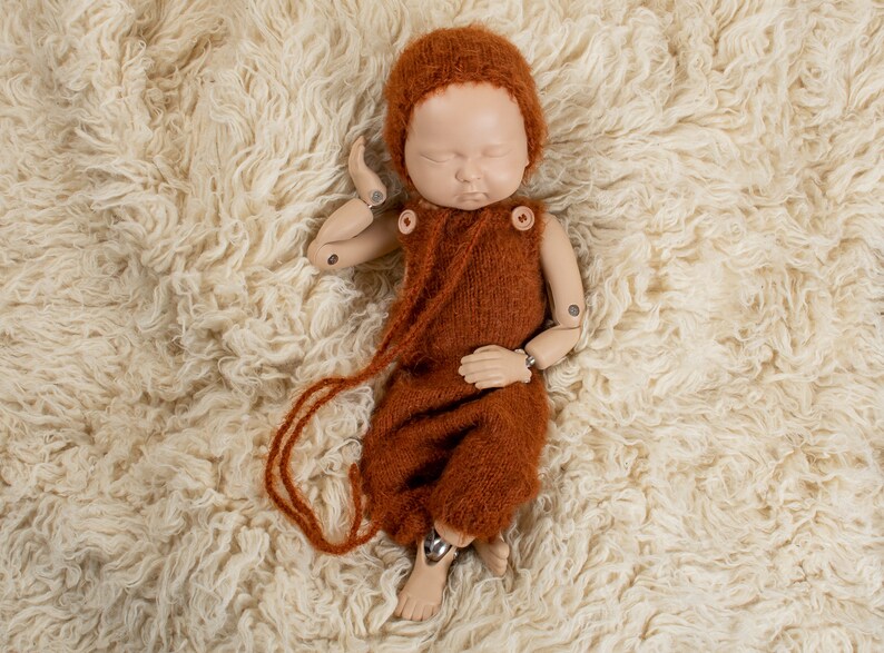 May include: A newborn photography prop featuring a doll wearing a rust-colored knitted romper and bonnet. The doll is lying on a cream-colored, fluffy rug. The romper has wooden button closures and a long, dangling tie.