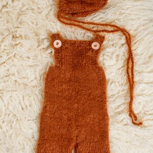 May include: A knitted rust-colored baby outfit set, including overalls with wooden buttons and a matching bonnet. The set is made of a soft, textured yarn and is displayed on a fluffy, cream-colored surface.