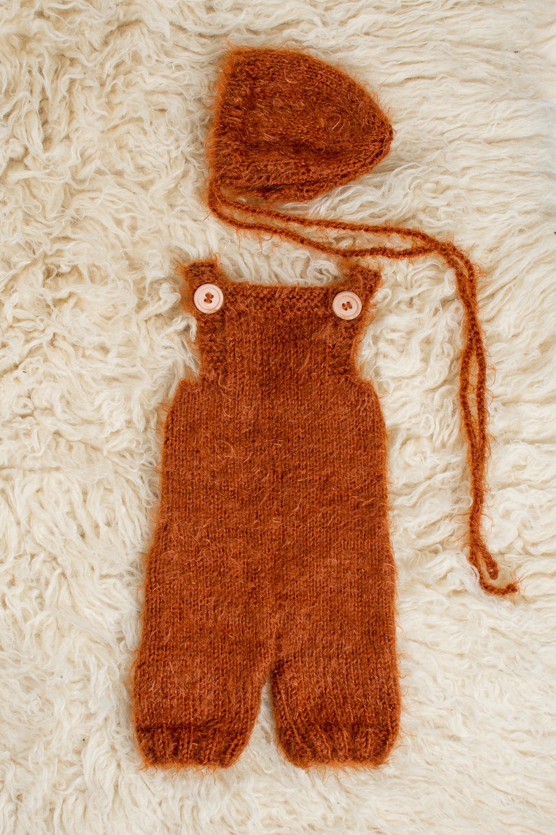 May include: A knitted rust-colored baby outfit set, including overalls with wooden buttons and a matching bonnet. The set is made of a soft, textured yarn and is displayed on a fluffy, cream-colored surface.