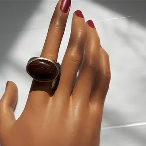 Peut inclure: Une bague en argent avec une pierre d'agate brune sertie dans un sertissage à bélière. La bague est sur le doigt d'un mannequin.