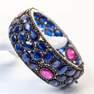May include: A silver-toned bracelet adorned with numerous oval-cut blue sapphires and a few pink gemstones. The gemstones are set closely together, creating a continuous pattern around the bracelet's band. The bracelet is displayed against a white background.