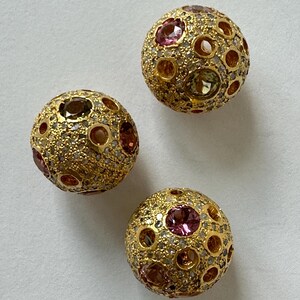May include: Three spherical beads with a textured gold-tone finish, each embedded with small clear pavé stones and larger, round, multi-colored faceted gemstones in shades of pink, orange, and yellow, arranged in a scattered, circular pattern.