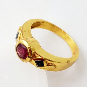 May include: A gold ring featuring a central round, bezel-set red gemstone. The band is accented with two small, square-cut black gemstones positioned on either side of the center stone, set within the polished gold metal surface of the ring.