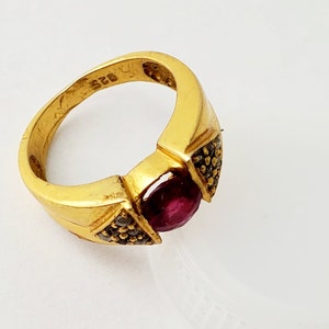 May include: A gold-toned ring featuring a central round-cut deep red gemstone. The band is marked with 925 on the interior. Triangular side settings on either side of the center stone are encrusted with small, dark, textured accent stones.