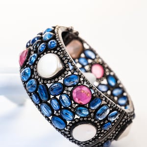May include: A silver bracelet adorned with a variety of gemstones. The bracelet features oval and round blue sapphires, pink tourmalines, and white moonstones. The stones are set in a silver band.