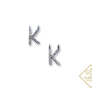 Peut inclure: Deux breloques en forme de lettre K argentée, chacune ornée d'une série de petites pierres claires et scintillantes incrustées sur la surface. Chaque breloque comporte deux petites boucles en haut. Un logo avec le texte BURHANI GEMS est visible.
