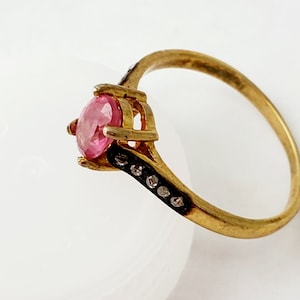 Peut inclure: Une bague en or avec une pierre précieuse rose taille ovale au centre, maintenue par quatre griffes. L'anneau présente un canal sombre et encastré sur le côté, orné d'une rangée de petites pierres claires rondes partant de la monture.