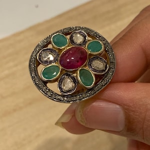 May include: An oval-shaped ring featuring a central ruby surrounded by emeralds and diamonds. The ring has a gold band and a detailed, textured setting. The gemstones are arranged in a floral pattern, creating a unique and elegant design.