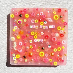 Puede incluir: Una pieza de arte cuadrada de resina rosa con elementos decorativos incrustados. La pieza presenta varias cuentas rosas, amarillas y doradas, estrellas y formas de conchas marinas. Las palabras "FEMININE RAGE" están escritas con cuentas de letras blancas.