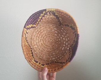 May include: A woven basket with a circular shape and a natural brown color. The basket has a purple and yellow rim.