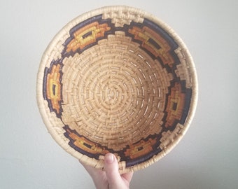 May include: A woven basket with a brown and orange geometric pattern around the rim. The basket is made of natural materials and has a circular shape.