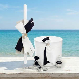 Puede incluir: Una vela blanca con cintas negras y blancas, una caja redonda blanca con una cinta negra y blanca y pequeños accesorios se exhiben sobre una mesa de madera blanca. El fondo presenta una playa con agua y cielo azules.