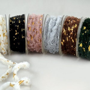 Op de afbeelding: Spoelen met decoratief garen in verschillende kleuren, waaronder wit met gouden sterren, zwart, roze, lichtblauw, bruin en donkergroen. Elke spoel is gevuld met pluizig garen en gouden folie accenten, perfect voor knutselprojecten.