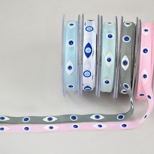 May include: Spools of ribbon in various colors, including light blue, white, mint green, gray, and pink. Each ribbon features a repeating pattern of blue and white evil eye designs. The ribbons are unspooled, showing the pattern.