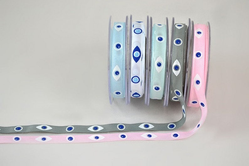 May include: Spools of ribbon in various colors, including light blue, white, mint green, gray, and pink. Each ribbon features a repeating pattern of blue and white evil eye designs. The ribbons are unspooled, showing the pattern.