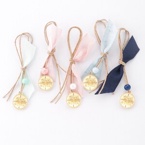 May include: Six gold charm favors with a cross design and the text "Jesus Saves" in Greek. Each charm is attached to a string with a small bead and a ribbon in a different color: light blue, pink, light green, navy blue, and two light blue.