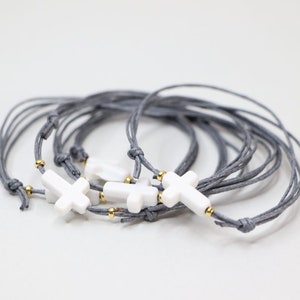 Könnte beinhalten: Ein Set aus grauen Schnurarmbändern mit weißen Kreuzanhängern und goldenen Perlen.