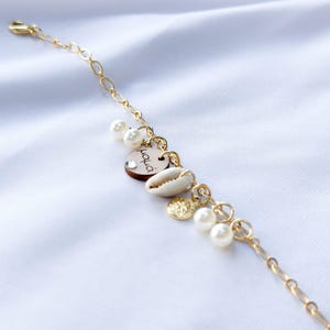 Peut inclure: Un bracelet en chaîne dorée avec un charme en bois qui dit "Juicy" et plusieurs charmes en perles blanches et en coquillages.