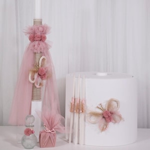 Puede incluir: Colección de elementos decorativos en rosa suave y blanco, que incluye una vela alta con detalles de tul y pompones, una pequeña caja de regalo, un frasco de perfume y una caja cilíndrica blanca con adornos florales. Dispuestos sobre una superficie blanca.