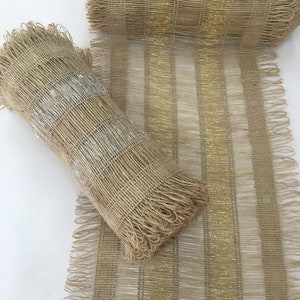 Op de afbeelding: Een rol beige jute lint met gouden en zilveren metallic accenten. Het lint heeft een gefranst randje en is geweven in een rasterpatroon.