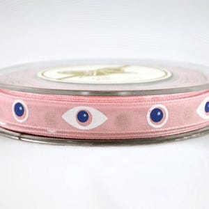 May include: A spool of pink ribbon with a repeating pattern of white-rimmed blue eye designs. The ribbon is wrapped around a clear plastic spool with a white label on top. The ribbon is approximately 1 inch wide.