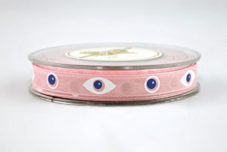 May include: A spool of pink ribbon with a repeating pattern of white-rimmed blue eye designs. The ribbon is wrapped around a clear plastic spool with a white label on top. The ribbon is approximately 1 inch wide.