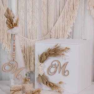 Puede incluir: Una caja blanca decorada con flores secas y una letra "M" de madera. La caja está adornada con una corona de plantas secas y una letra "M" de madera. También hay un colgante decorativo con la letra "M" y una pequeña botella de vidrio.