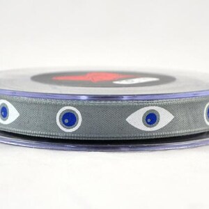 May include: A spool of gray ribbon with a repeating eye pattern in white and blue. The ribbon is wound on a clear plastic spool with a black and red design on top.