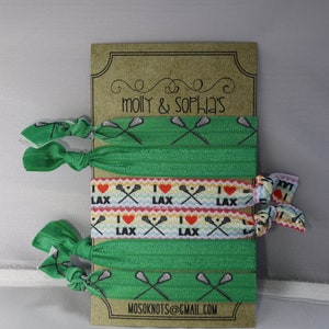 May include: A set of six green and white elastic hair ties with lacrosse stick and heart designs. The hair ties are packaged on a brown card with the text "molly & sophia's" and "mosoknots@gmail.com".