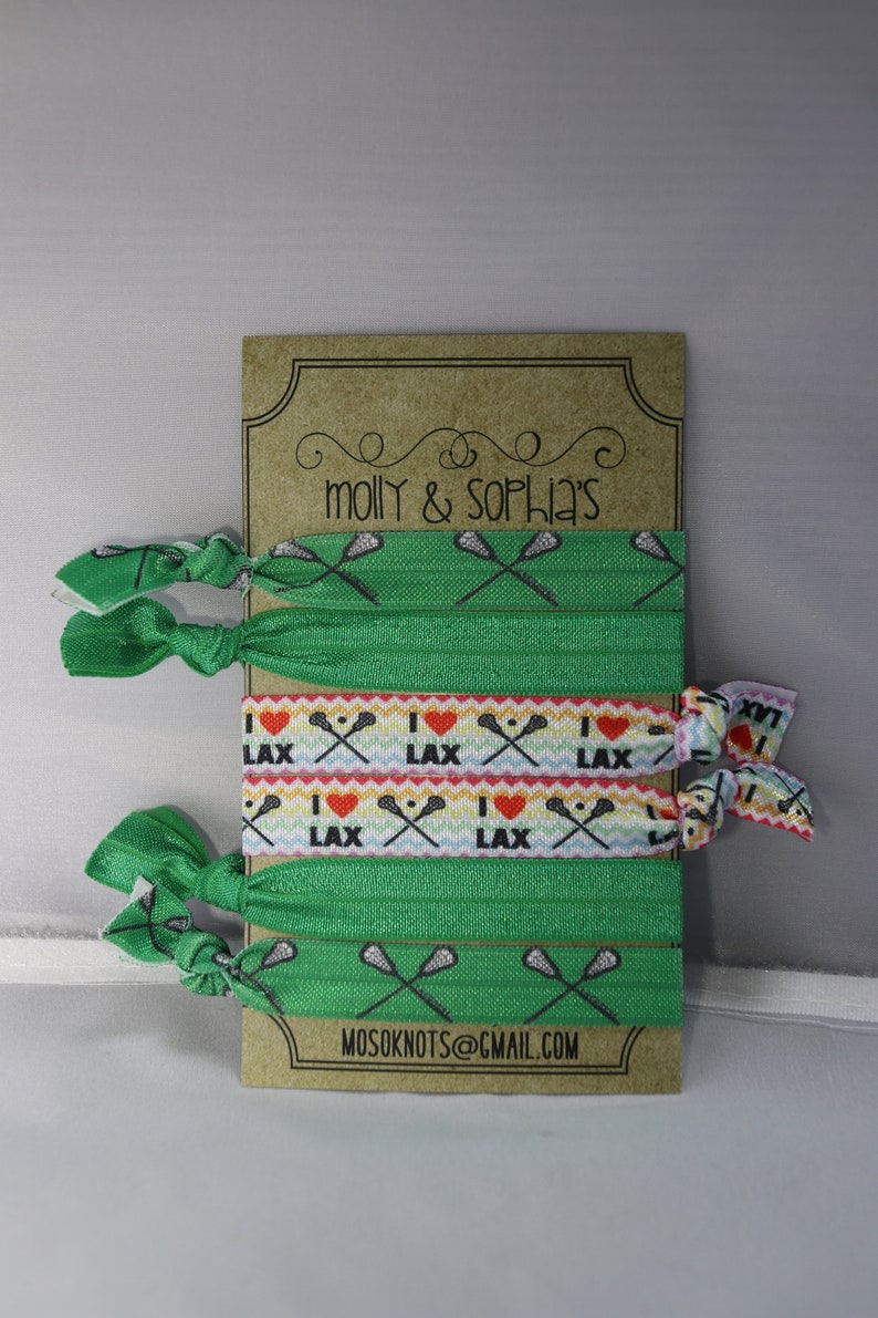 May include: A set of six green and white elastic hair ties with lacrosse stick and heart designs. The hair ties are packaged on a brown card with the text "molly & sophia's" and "mosoknots@gmail.com".