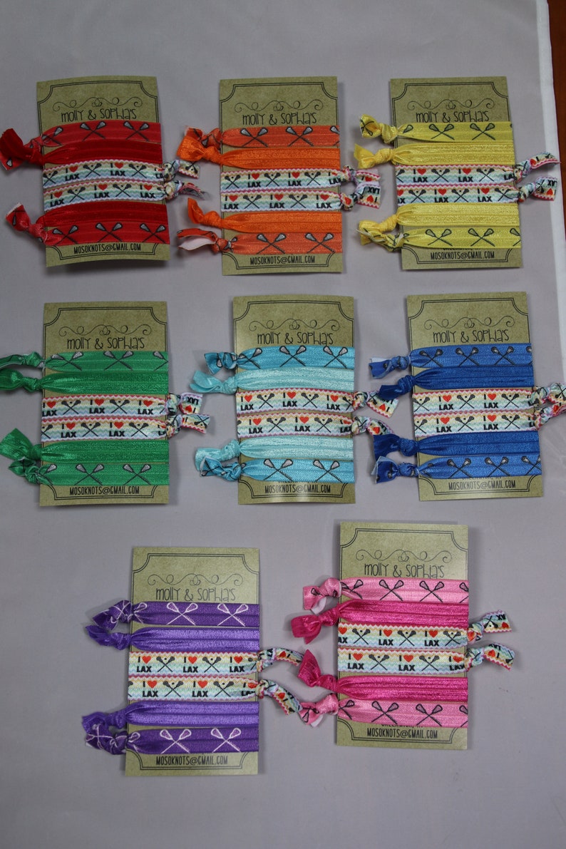 May include: Set of ten colorful hair scrunchies with a lacrosse theme. The scrunchies are made of elastic fabric and have a printed design with lacrosse sticks and the words "I Love Lax". The scrunchies are packaged on individual cards with the name "Molly & Sophia's" and an email address.