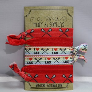 May include: A set of six red and white elastic hair ties with a lacrosse stick and "I love LAX" design. The hair ties are displayed on a brown card with the text "molly & sophia's" and "mosoknots@gmail.com".