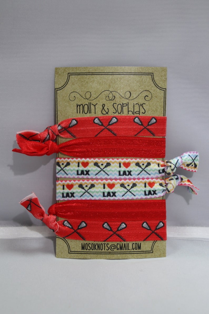 May include: A set of six red and white elastic hair ties with a lacrosse stick and "I love LAX" design. The hair ties are displayed on a brown card with the text "molly & sophia's" and "mosoknots@gmail.com".