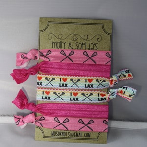 May include: A set of six pink and white hair ties with a lacrosse stick and heart design. The hair ties are packaged on a brown card with the text "molly & sophia's" and "mosoknots@gmail.com".